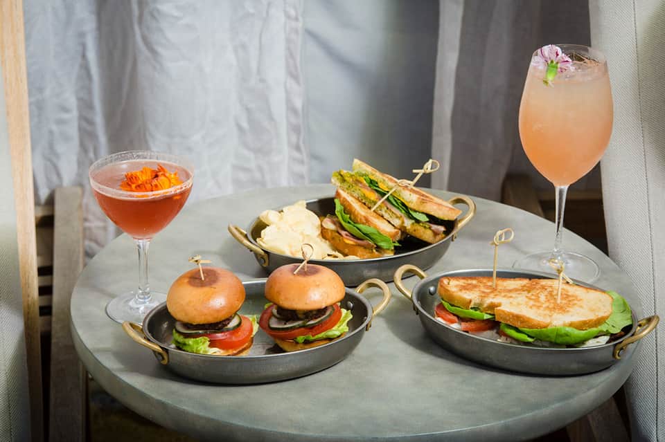 Gourmet appetizers and craft cocktails on a concrete table including sliders, sandwiches, and colorful beverages