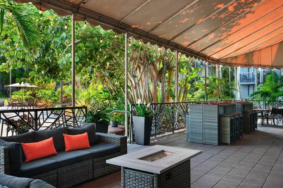 Covered outdoor lounge area with black seating, red cushions, fire pit table, and lush tropical plants