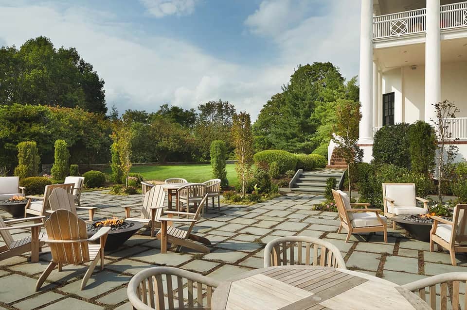 Elegant outdoor patio with Adirondack chairs, stone pathways, manicured hedges, and white colonial mansion