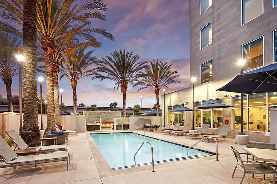 Illuminated resort pool at dusk with palm trees, lounge chairs, and modern building with lit windows