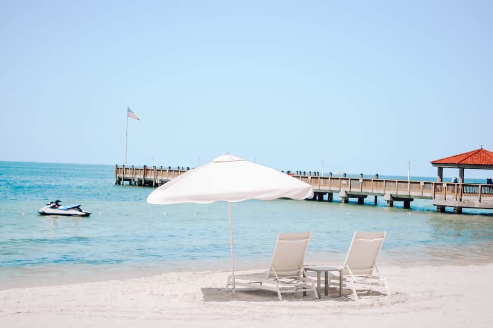 Beachfront lounge chairs with white umbrella overlooking turquoise water and wooden pier