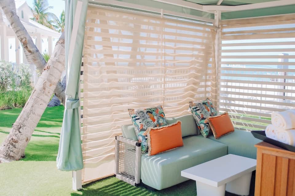 Covered outdoor patio with mint green seating, striped privacy screens, colorful throw pillows, and ocean view
