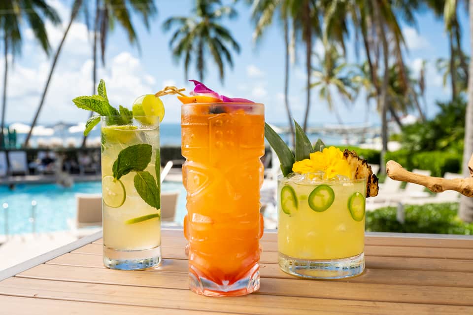 Three tropical cocktails on a wooden deck with palm trees and pool in background