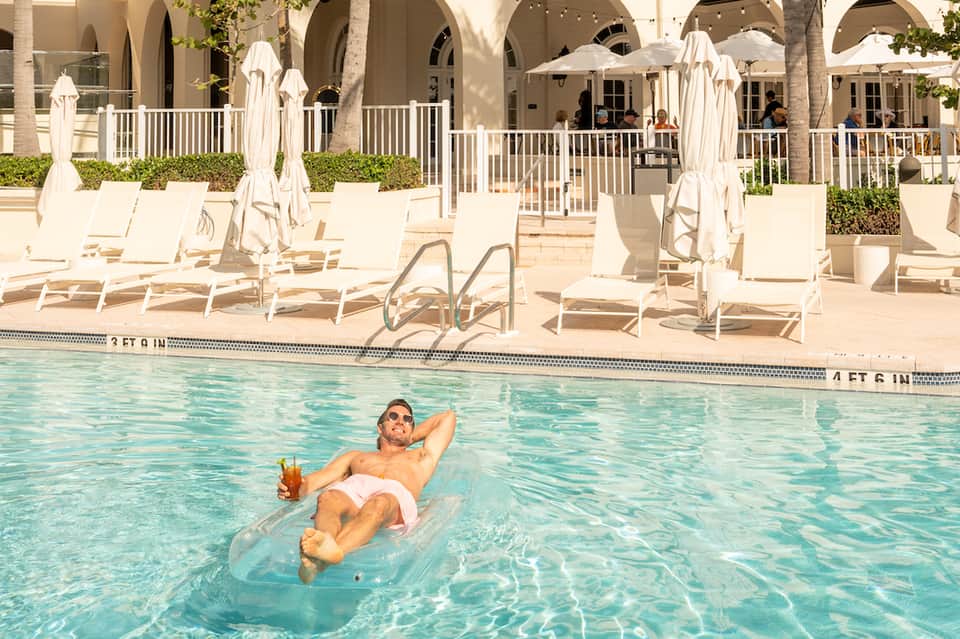 Man floating on a raft in a crystal-clear pool with lounge chairs and white umbrellas on the terrace