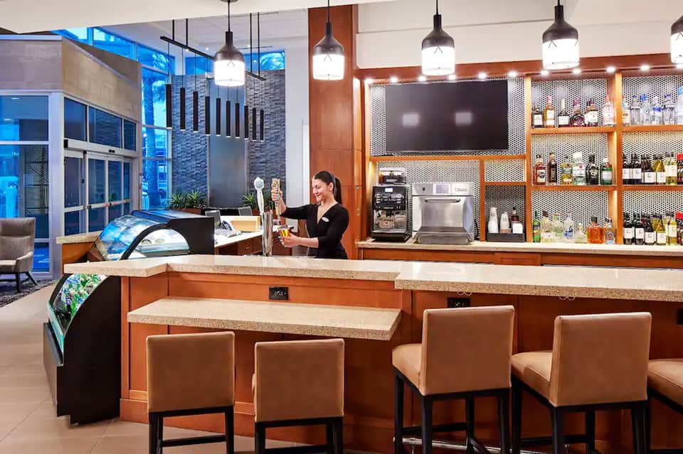 Modern hotel bar with orange wood cabinetry, pendant lighting, and stocked shelves behind counter