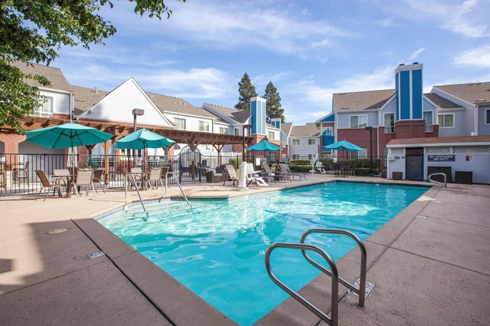 Resort swimming pool with turquoise water, lounge chairs under teal umbrellas, and residential buildings in background