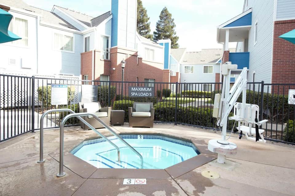 Outdoor spa hot tub surrounded by black fencing with residential buildings and pine trees in background