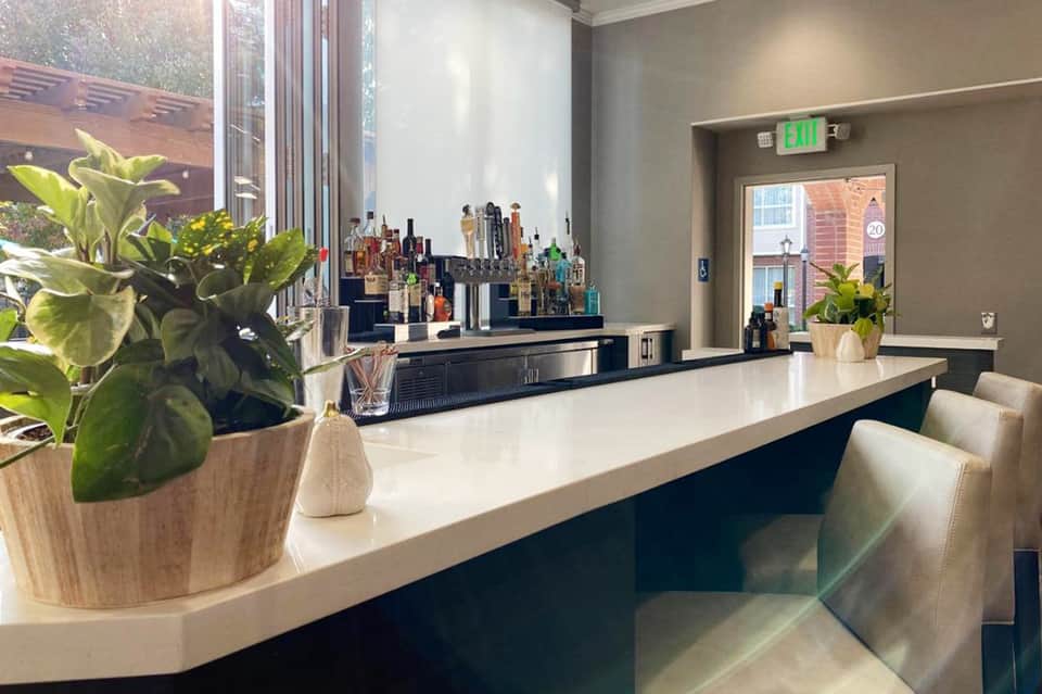 Modern hotel bar with white counter, potted plants, and extensive liquor selection behind glass