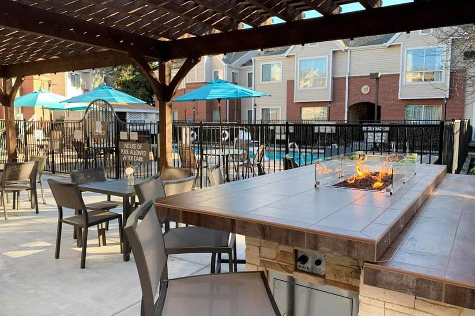 Outdoor patio with fire pit table, blue umbrellas, wooden pergola, and residential buildings in background