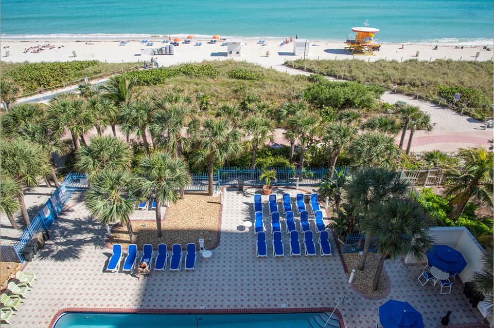 Aerial view of beach with blue umbrellas, palm trees, lifeguard stand, and turquoise ocean water