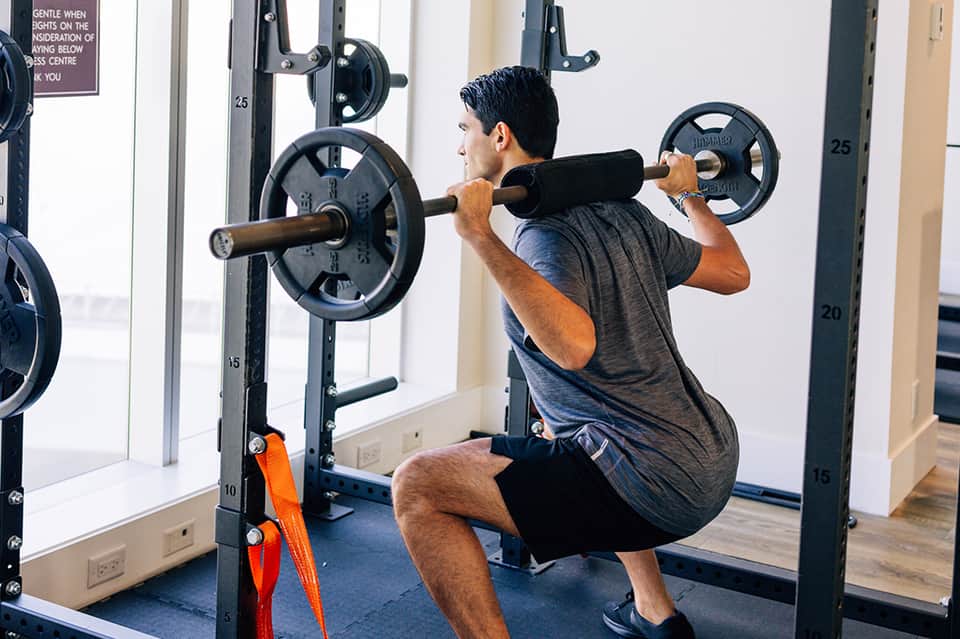 Man performing barbell squat exercise in gym with weight plates and mirrors