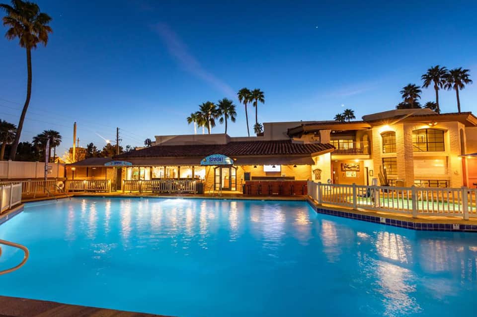 Illuminated resort pool area at dusk with yellow building, Splash bar, and palm trees lining the water