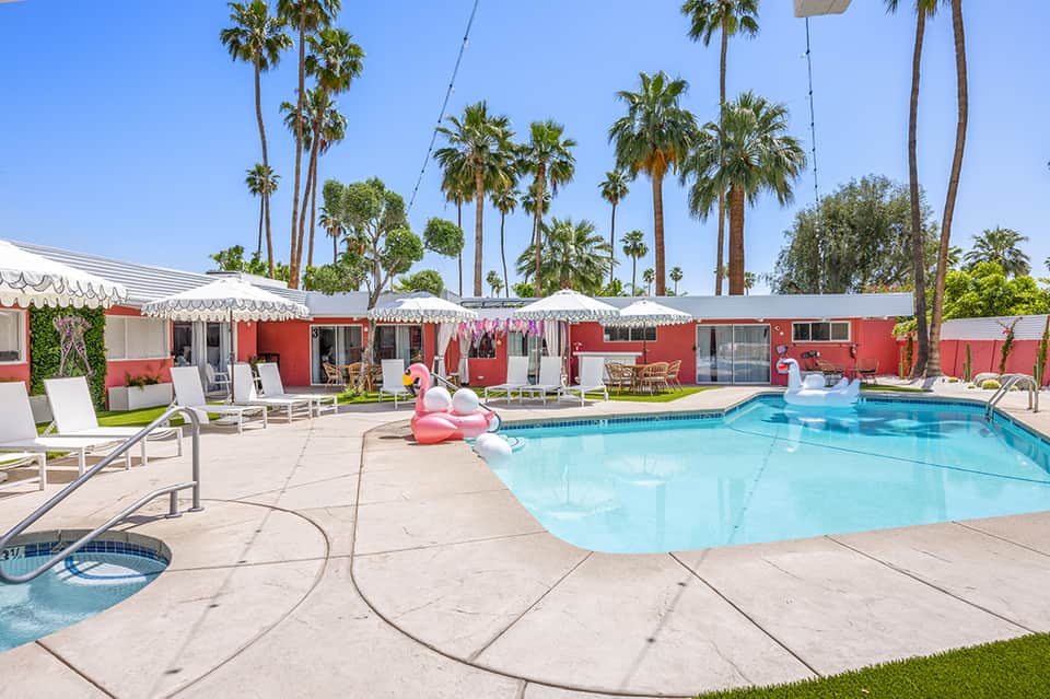 Retro mid-century motel with red buildings, palm trees, swimming pool, and lounge chairs under white umbrellas
