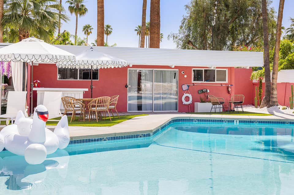 Red mid-century modern house with pool, white umbrella, and lounge chairs surrounded by palm trees