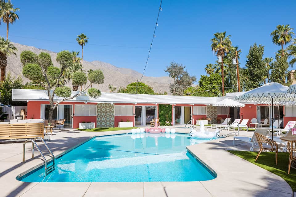 Retro-style resort pool surrounded by red and white mid-century buildings with palm trees and desert mountains in background