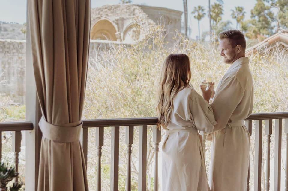 Couple in white robes on a balcony overlooking desert landscape with mountains and palm trees