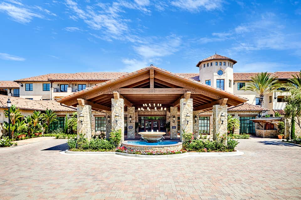 Luxury resort entrance with stone columns, wooden beams, central fountain, and manicured landscaping under blue sky