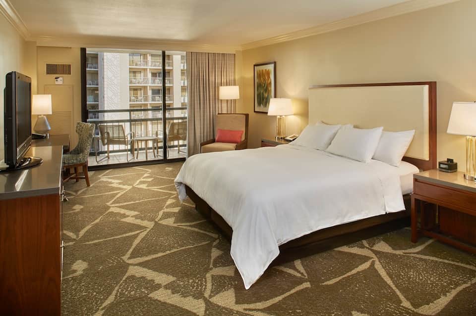 Spacious hotel bedroom with king bed, cream bedding, wood furnishings, and balcony overlooking city buildings