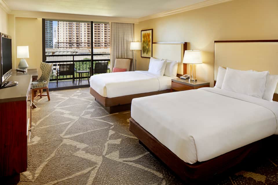 Hotel room with two queen beds, balcony view of high-rise buildings, warm beige decor