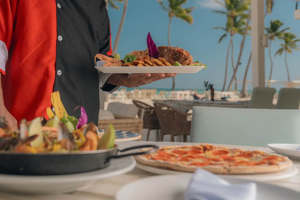 Server presenting appetizers and pizza at beachfront restaurant with ocean view and palm trees