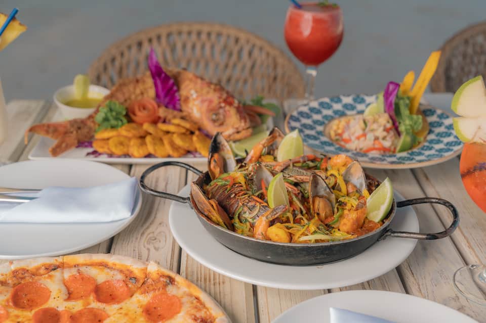 Beachfront dining table with seafood paella, grilled fish, and cocktails with colorful garnishes