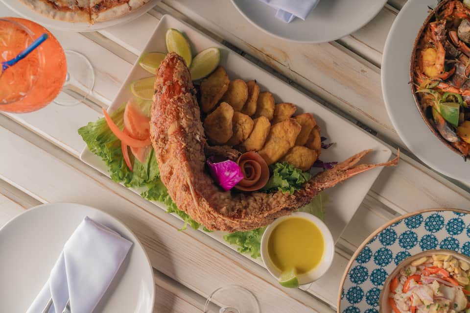 Overhead view of grilled whole fish with fried plantains, lime wedges, and seafood appetizers