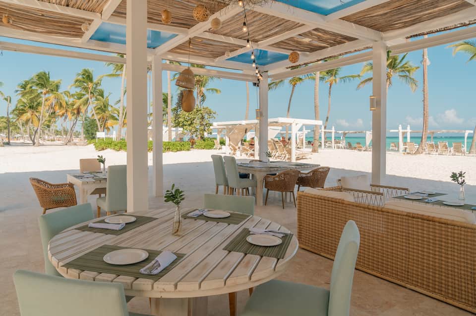 Beachfront dining pavilion with white pergola, striped tables, wicker chairs, and turquoise ocean view