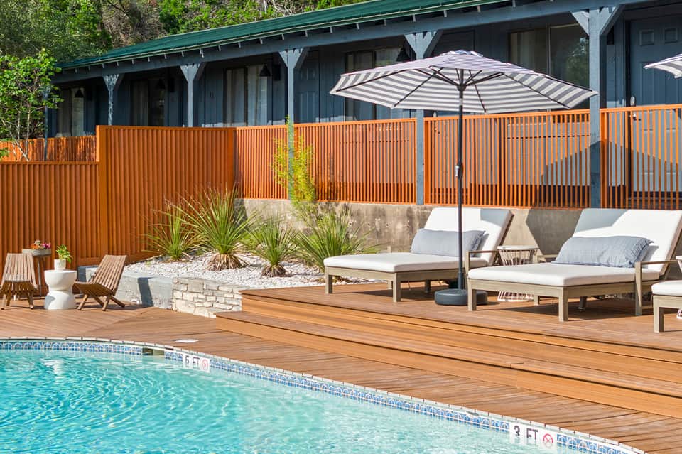 Resort pool deck with wooden loungers, striped umbrella, modern house with orange railing, and lush landscaping