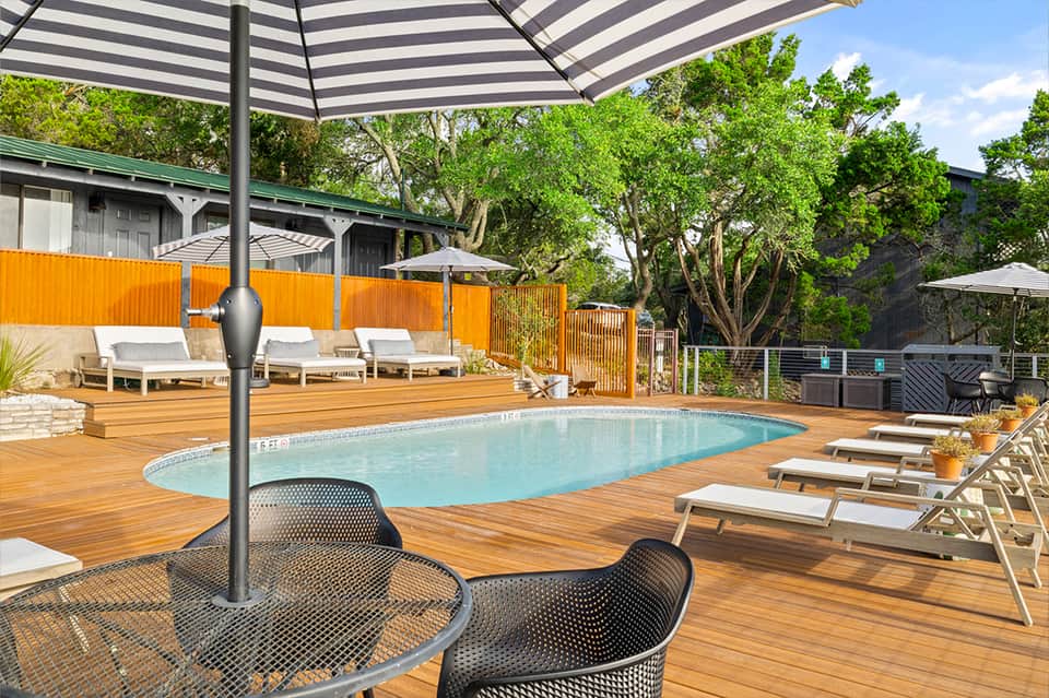 Resort pool deck with turquoise water, wooden decking, striped umbrellas, lounge chairs, and orange privacy fence