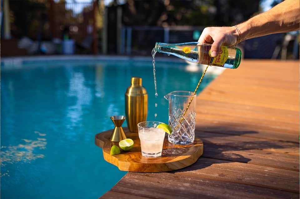 Hand pouring beverage at poolside serving station with cocktail glasses and tropical setting