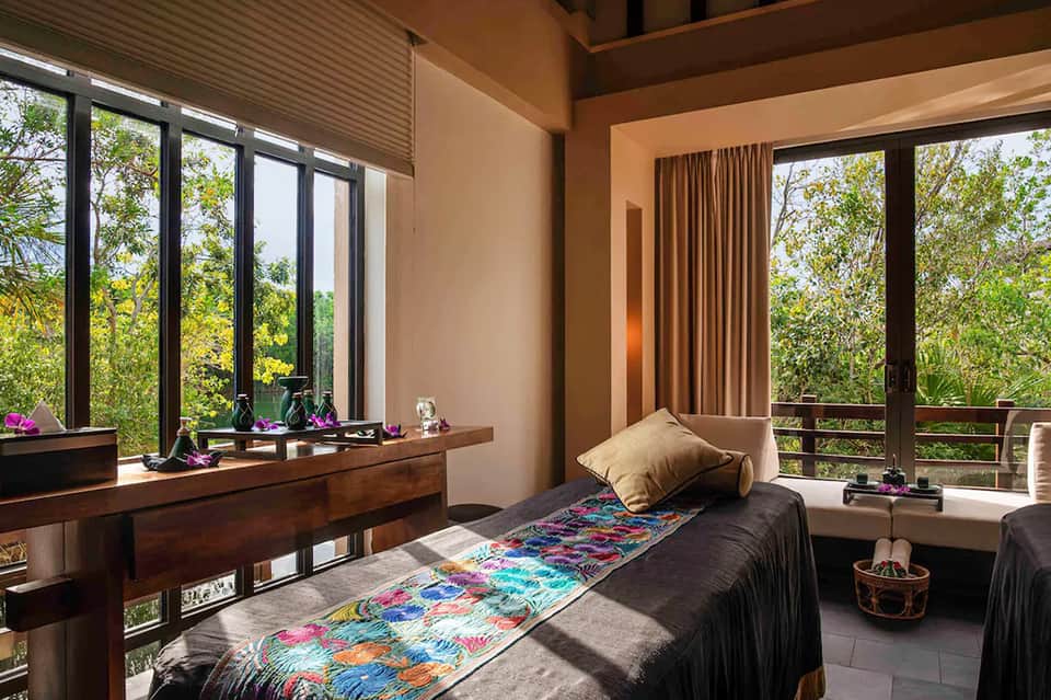 Modern bedroom with black bed, colorful throw blanket, wooden desk, and floor-to-ceiling windows overlooking tropical forest