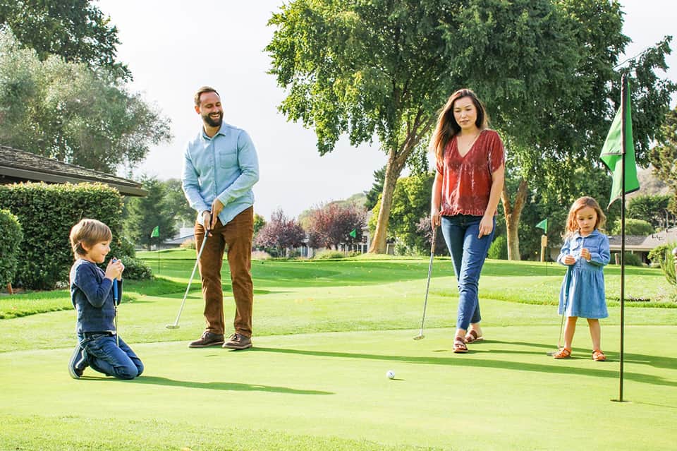 Family playing miniature golf on a manicured green lawn with trees and a flag in background
