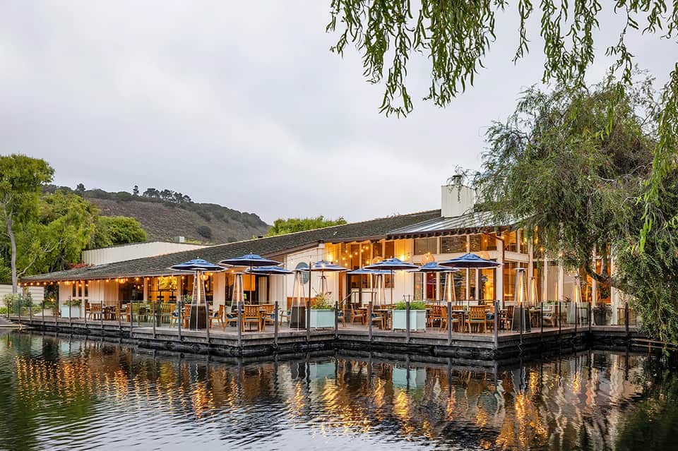 Modern waterfront restaurant with wooden deck, large windows, and blue umbrellas overlooking a river