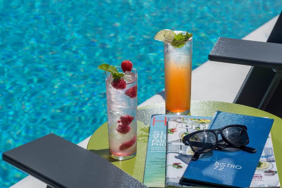 Poolside cocktails with fresh berries and citrus garnish, sunglasses, and magazines on table overlooking blue water