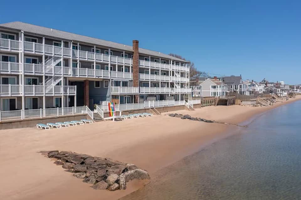 Beachfront hotel with white railings and beach loungers on sandy shore beside calm water