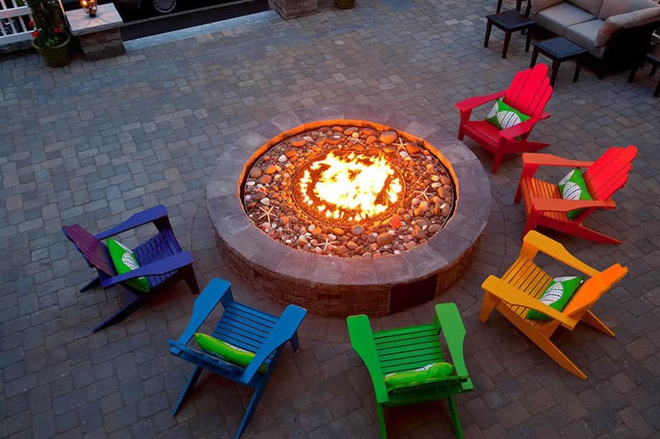 Colorful Adirondack chairs surrounding an illuminated circular fire pit on a brick patio at dusk