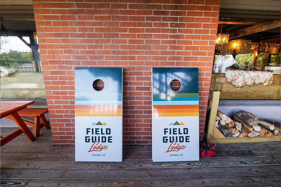 Two cornhole game boards with Field Guide Lodge branding displayed on brick patio
