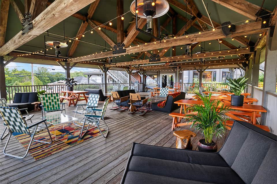 Covered outdoor deck with wooden beams, string lights, colorful lounge chairs, and orange dining tables overlooking green landscape