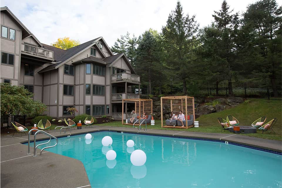 Resort pool with white floating decor, modern lounge chairs, wooden pergolas, and multi-story house