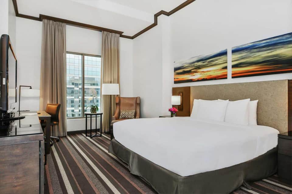 Modern hotel bedroom with king bed, work desk, and floor-to-ceiling windows with city views