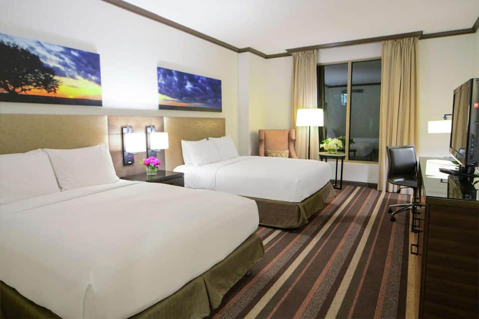Hotel room with two queen beds, tan headboards, artwork, desk area, and balcony with curtains