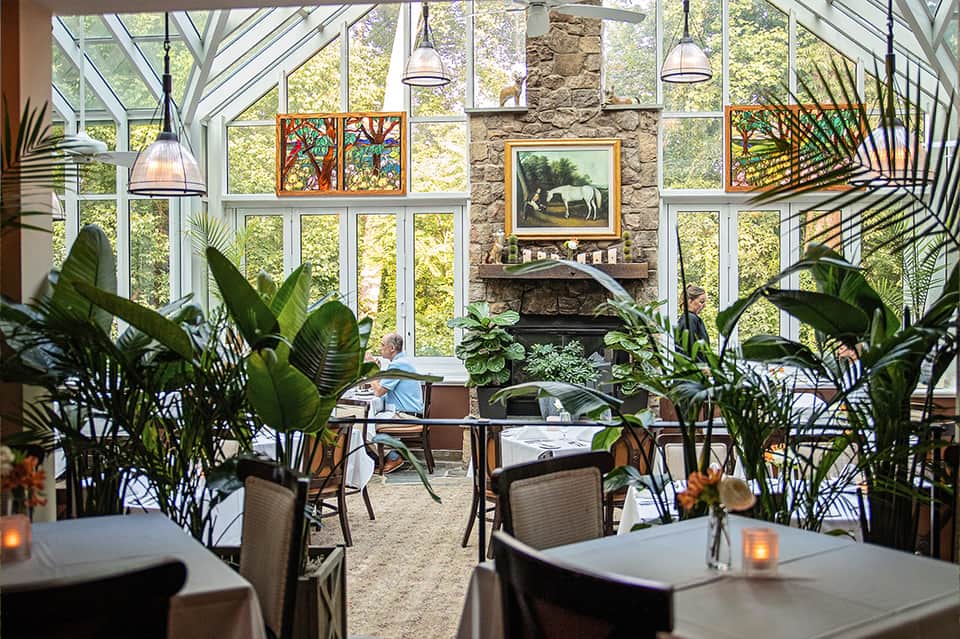 Sunlit conservatory dining area with stone fireplace, plants, and large glass windows
