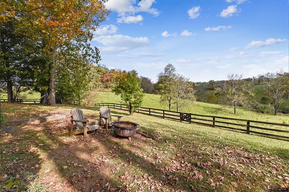Pastoral landscape with two Adirondack chairs by fire pit, wooden fencing, and rolling green fields