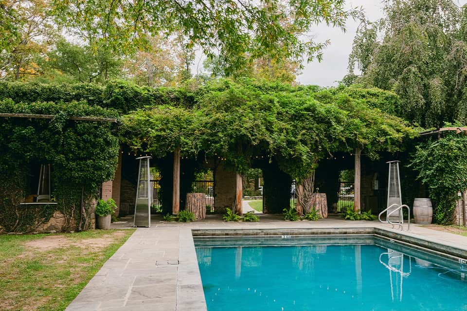 Resort swimming pool with ivy-covered pergola structure and lounge chairs under mature trees
