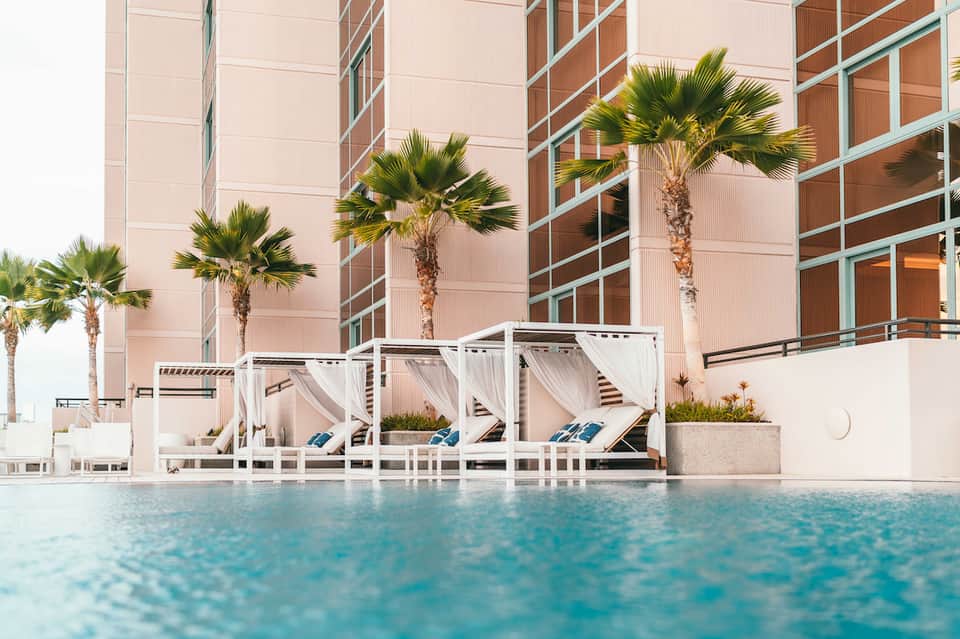 Resort pool with white cabanas, palm trees, modern pink building facade with green accents