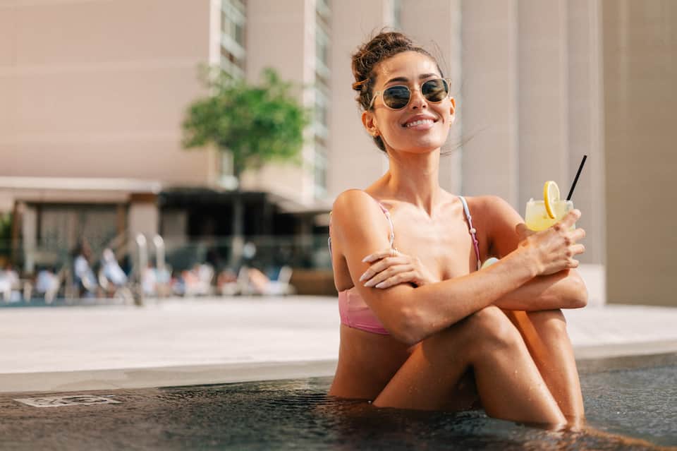 Woman in pink bikini sitting in shallow water holding a lemon drink, wearing sunglasses