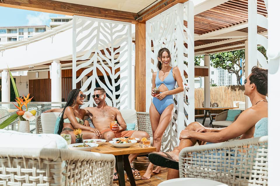 Resort pool cabana with people dining, featuring white geometric privacy screens and wooden pergolas