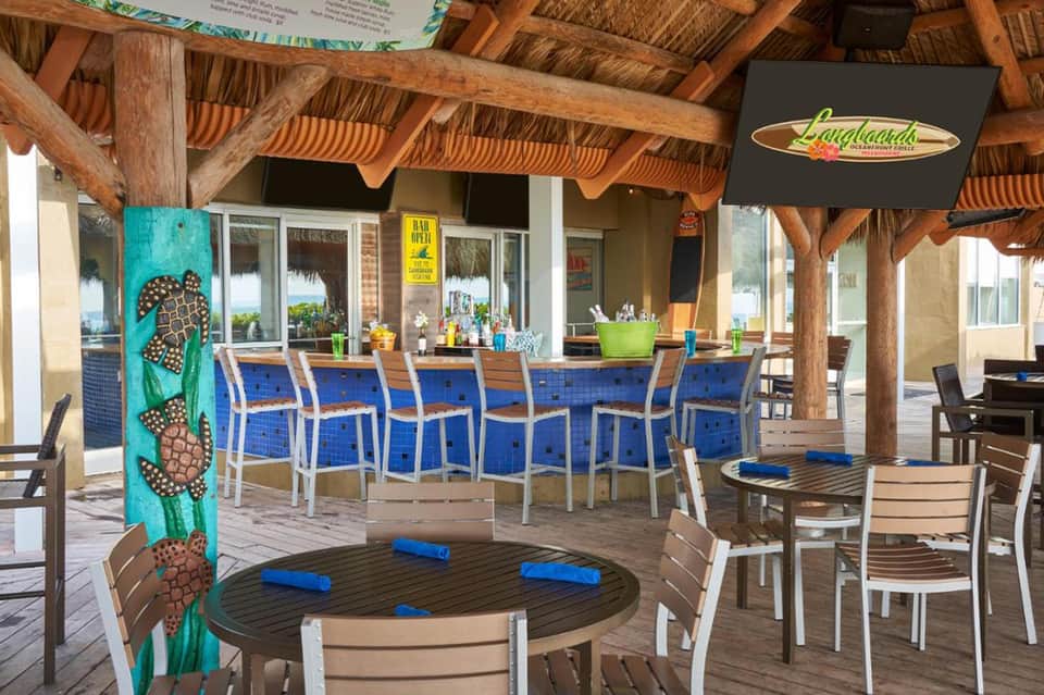 Tropical beachfront bar with thatched roof, blue cushioned seating, and wooden tables under covered patio