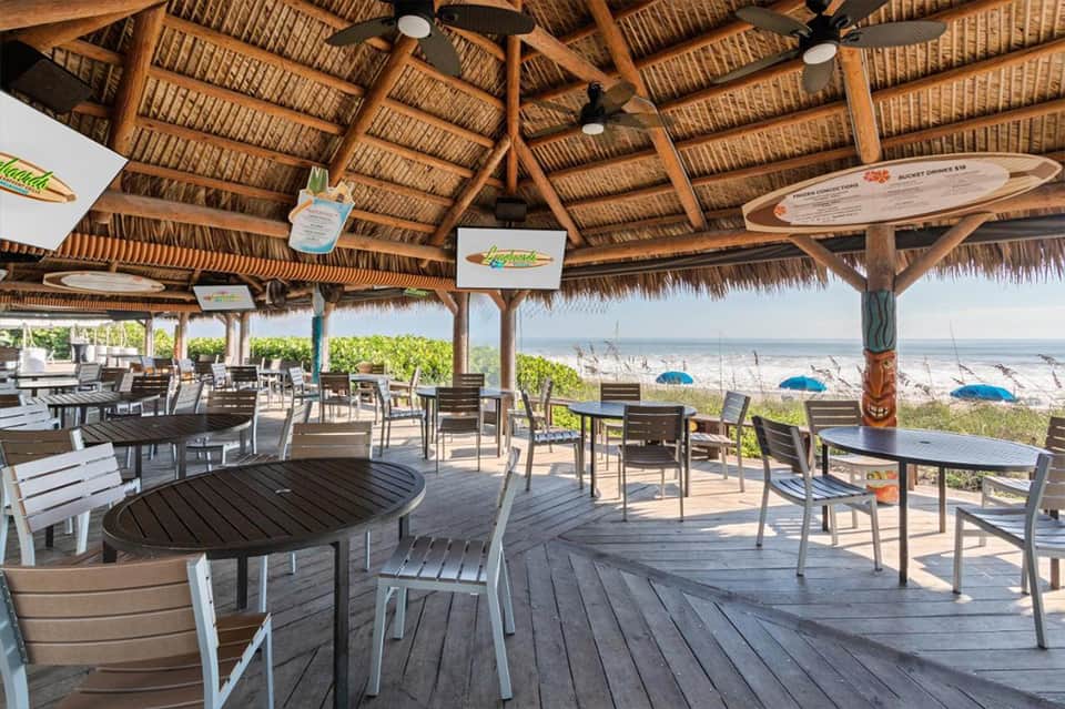 Beachfront tiki bar with wooden thatched roof, dining tables, ocean view, and colorful umbrellas on sand