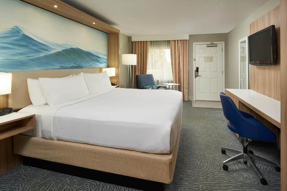 Modern hotel bedroom with queen bed, mountain artwork, work desk, flat-screen TV, and neutral earth-tone decor